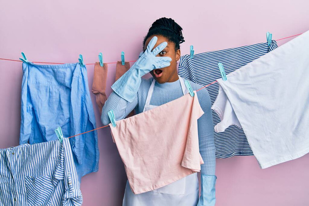 African american woman with braided hair washing clothes at clothesline peeking in shock covering face and eyes with hand, looking through fingers with embarrassed expression.  - Foto, Imagen