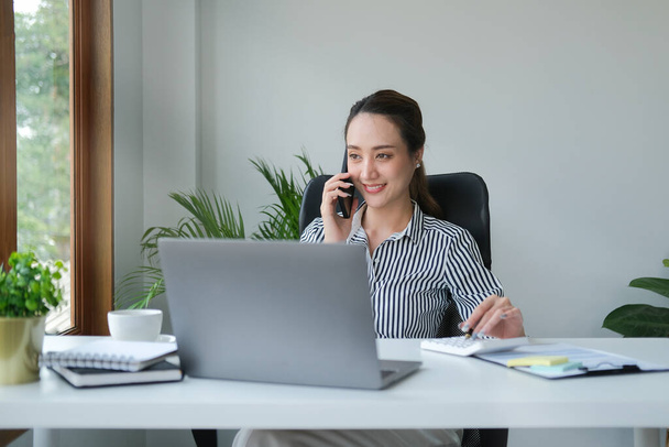 Urocza młoda azjatycka bizneswoman siedzi w jasnym nowoczesnym biurze i rozmawia przez telefon komórkowy. - Zdjęcie, obraz