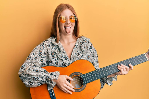 Nuori irlantilainen nainen yllään boho tyyli pelaa klassista kitaraa törröttää kielen ulos onnellinen hauska ilme.  - Valokuva, kuva