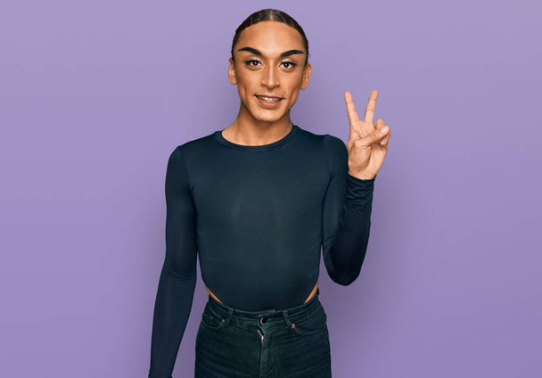 Der hispanische Transgender-Mann mit Make-up und langen Haaren in lässiger Kleidung lächelt mit glücklichem Gesicht in die Kamera und macht ein Siegeszeichen. Nummer zwei.  - Foto, Bild