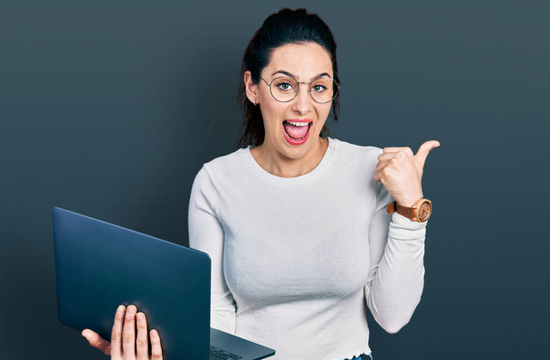 Молода іспаномовна жінка працює за допомогою комп'ютерного ноутбука, вказуючи великий палець в сторону посміхаючись щасливий з відкритим ротом
  - Фото, зображення