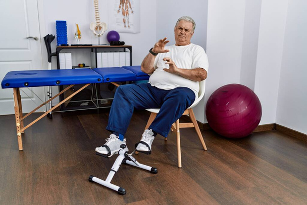 Старший кавказький чоловік у фізіотерапевтичній клініці використовує педальну вправу огидний вираз, незадоволений і боїться робити огидне обличчя, тому що реакція відрази. з піднятими руками
  - Фото, зображення