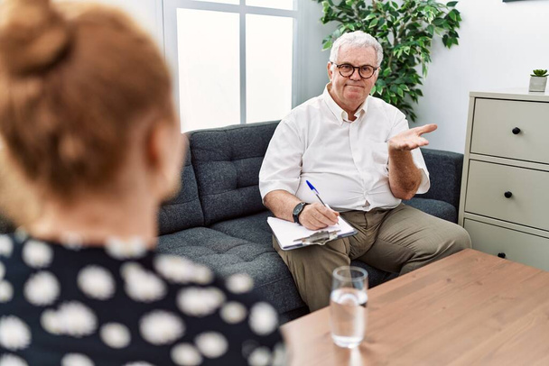 Senior psycholog muž v konzultační kanceláři ukazuje stranou s rukama otevřenýma dlaněmi ukazující kopírovací prostor, prezentující reklamu s nadšeným úsměvem  - Fotografie, Obrázek