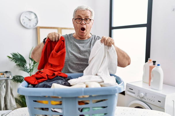Senior caucasian man doing laundry holding clothes from wicker basket afraid and shocked with surprise and amazed expression, fear and excited face.  - Zdjęcie, obraz