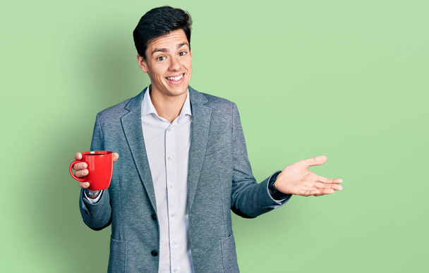 Junger hispanischer Geschäftsmann im Business-Stil trinkt eine Tasse Kaffee und zelebriert seine Leistung mit glücklichem Lächeln und Siegerausdruck mit erhobener Hand  - Foto, Bild