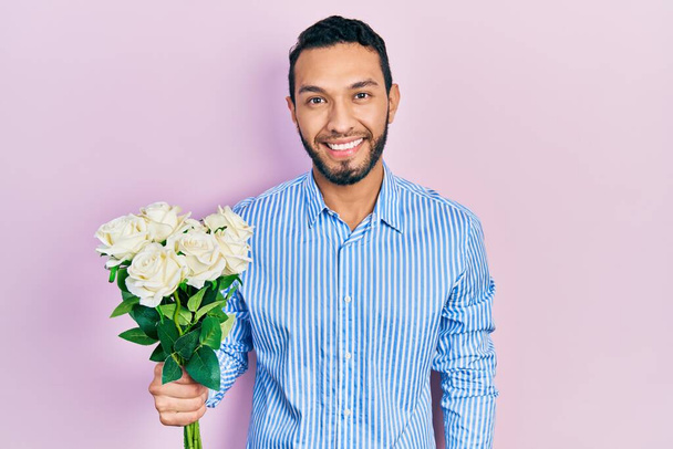 Латиноамериканец с бородой, держа букет белых цветов, смотрит позитивно и счастливо стоя и улыбаясь с уверенной улыбкой, показывая зубы  - Фото, изображение
