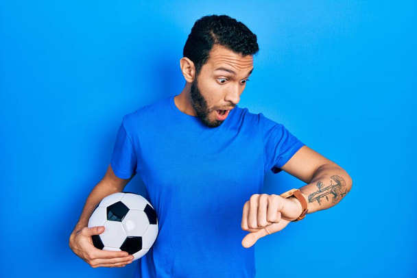 Homme hispanique avec barbe tenant ballon de football regardant l'heure de la montre inquiet, peur de se faire tard  - Photo, image