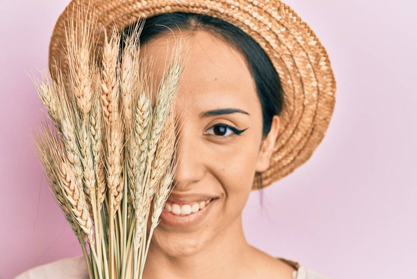Joven chica hispana con sombrero de verano sosteniendo trigo espiga sobre el ojo sonriendo con una sonrisa feliz y fresca en la cara. mostrando dientes.  - Foto, Imagen