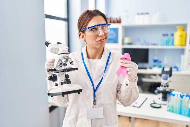 Młoda brunetka pracuje w laboratorium naukowca trzymając różową wstążkę uśmiechając się patrząc w bok i patrząc myśląc.  - Zdjęcie, obraz