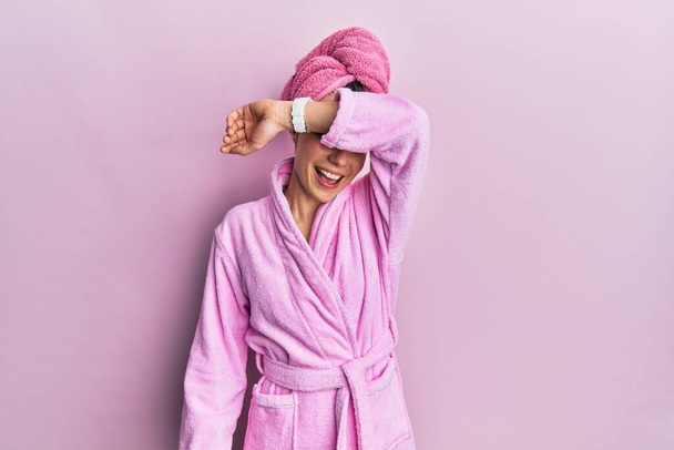 Young blonde woman wearing shower towel cap and bathrobe covering eyes with arm smiling cheerful and funny. blind concept.  - 写真・画像