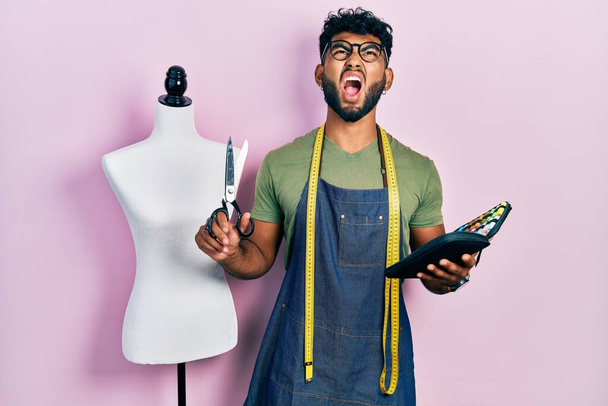Arabische man met baard dressmaker ontwerper vasthouden schaar en naaien kit boos en gek schreeuwen gefrustreerd en woedend, schreeuwen van woede opkijken.  - Foto, afbeelding