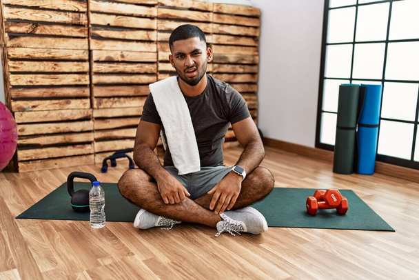 Joven hombre indio sentado en la alfombra de entrenamiento en el gimnasio en la cara de choque, mirando escéptico y sarcástico, sorprendido con la boca abierta  - Foto, Imagen