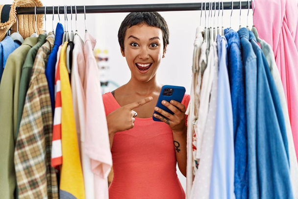 Young hispanic woman with short hair searching clothes on clothing rack using smartphone smiling happy pointing with hand and finger  - Φωτογραφία, εικόνα