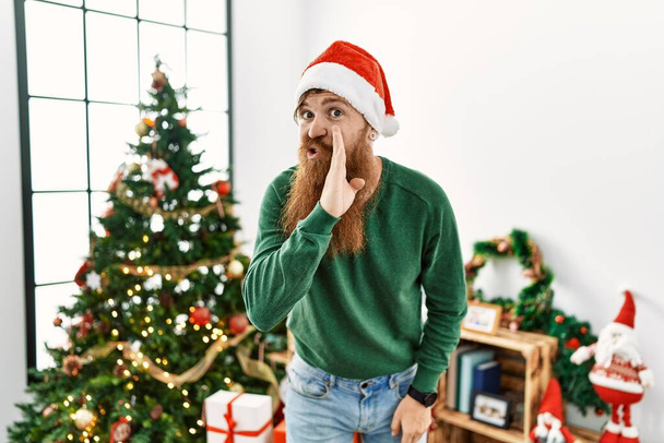 Homme rousse à longue barbe portant un chapeau de Noël par arbre de Noël main sur la bouche racontant une rumeur secrète, chuchotant conversation malicieuse  - Photo, image