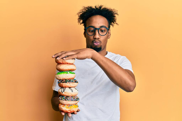 Jovem afro-americano com barba segurando pilha de saborosos donuts coloridos fazendo cara de peixe com boca e olhos espremendo, louco e cômico.  - Foto, Imagem