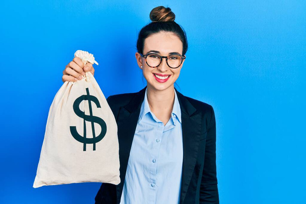 Young hispanic girl wearing business clothes holding dollars bag looking positive and happy standing and smiling with a confident smile showing teeth  - Foto, afbeelding
