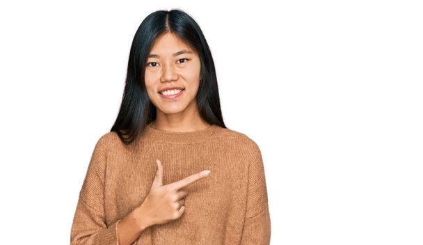 Kaunis nuori aasialainen nainen yllään rento talvi villapaita iloinen hymyillen kasvot osoittaa kädellä ja sormella sivulle onnellinen ja luonnollinen ilme kasvoilla  - Valokuva, kuva