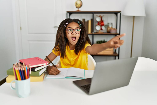 Young african american girl doing homework at home pointing with finger surprised ahead, open mouth amazed expression, something on the front  - 写真・画像