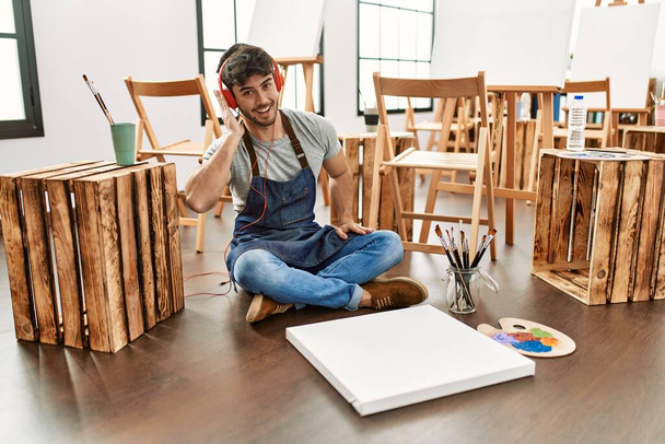 Mladý hispánský umělec muž se šťastně usmívá a poslouchá hudbu malování v ateliéru umění. - Fotografie, Obrázek