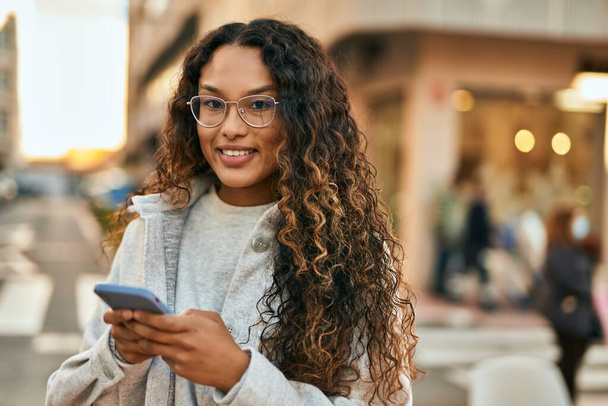 Jeune femme latine souriant heureux en utilisant un smartphone à la ville. - Photo, image