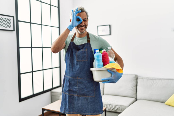 Middle age hispanic man wearing cleaner apron holding cleaning products smiling happy doing ok sign with hand on eye looking through fingers  - Fotó, kép