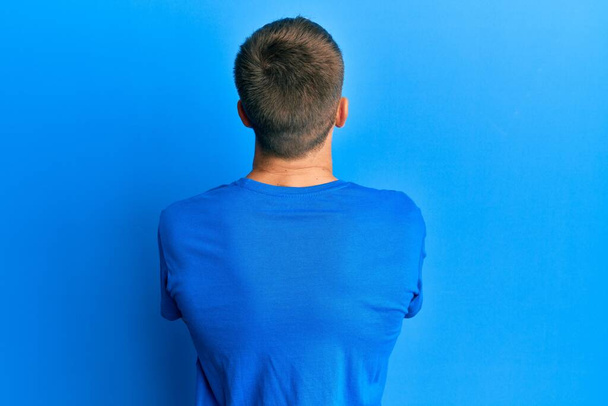 Νεαρός καυκάσιος με μπλε μπλουζάκι που στέκεται πίσω κοιτώντας αλλού με σταυρωμένα χέρια.  - Φωτογραφία, εικόνα