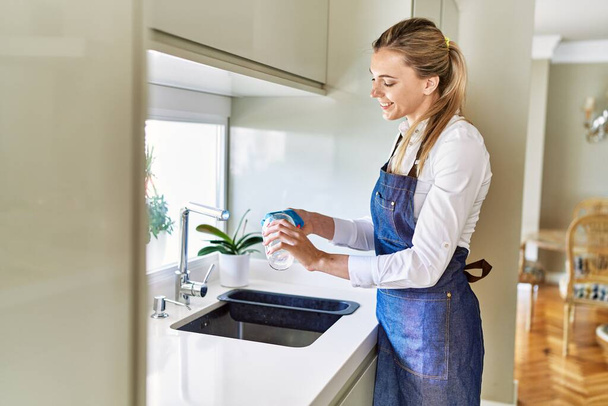 Giovane donna bionda sorridente vetro di lavaggio fiducioso in cucina - Foto, immagini