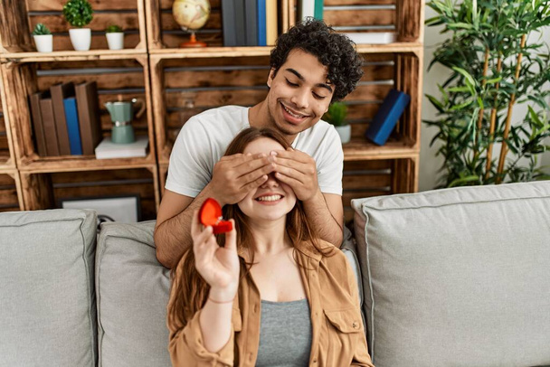 Молодая пара улыбается счастливо сидя на диване держа обручальное кольцо дома. - Фото, изображение
