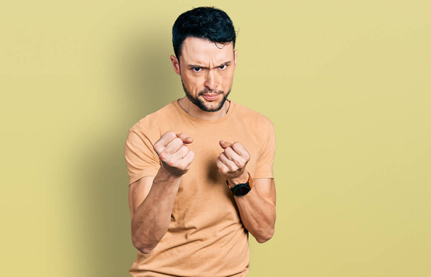 Латиноамериканець з бородою у повсякденній футболці готовий боротися з жестом захисту кулаками, сердитим і засмученим обличчям, побоюючись проблем.  - Фото, зображення