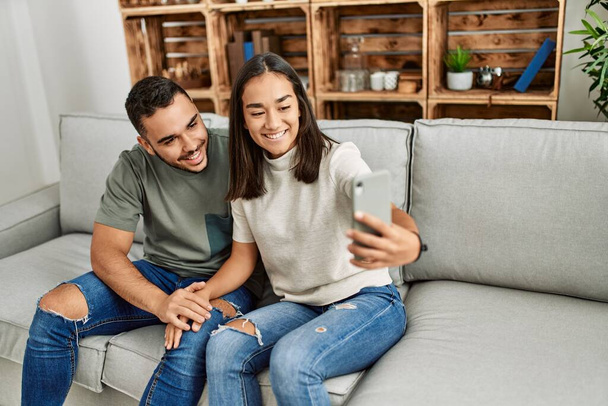 Joven pareja latina sonriendo feliz haciendo selfie junto al smartphone en casa. - Foto, imagen