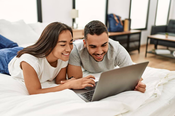 Joven pareja latina sonriendo feliz usando portátil en el dormitorio - Foto, imagen