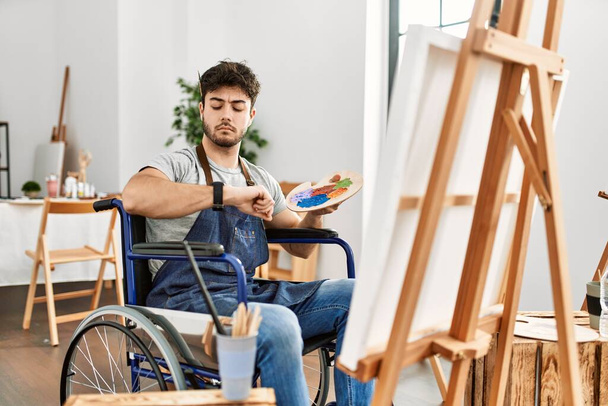 Junger hispanischer Mann sitzt auf Rollstuhlmalerei im Kunststudio und überprüft die Uhrzeit an der Armbanduhr, entspannt und selbstbewusst  - Foto, Bild