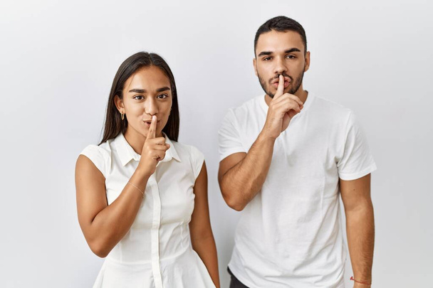 Young interracial couple standing together in love over isolated background asking to be quiet with finger on lips. silence and secret concept.  - Photo, image