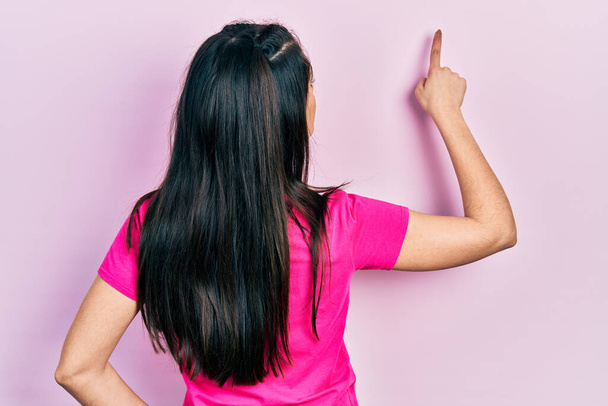 Menina hispânica jovem vestindo camisa casual rosa t posando para trás apontando para a frente com a mão do dedo  - Foto, Imagem