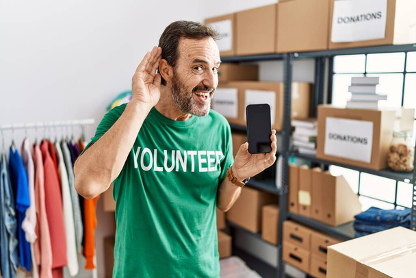 Uomo di mezza età con la barba che indossa una t-shirt volontaria che tiene lo smartphone sorridente con le mani sull'orecchio ascoltando voci o pettegolezzi. concetto di sordità.  - Foto, immagini