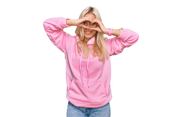 Young blonde woman wearing casual sweatshirt doing heart shape with hand and fingers smiling looking through sign  - Fotoğraf, Görsel