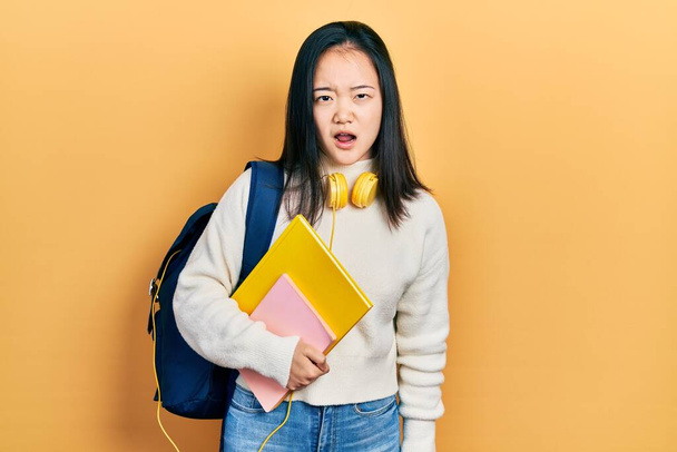 Giovane ragazza cinese tenendo zaino studente e libri in faccia shock, cercando scettico e sarcastico, sorpreso con la bocca aperta  - Foto, immagini
