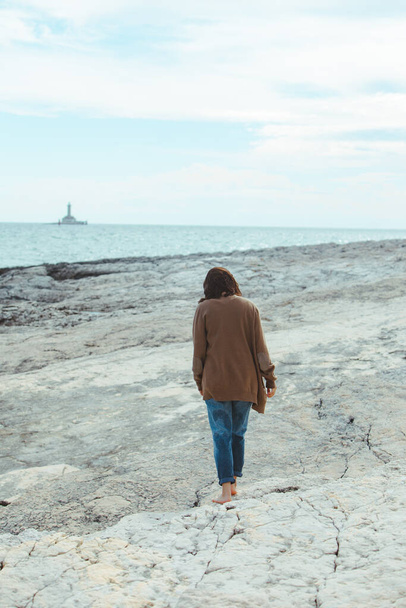 Nainen kävelee kivinen meren rannalla märät farkut majakka taustalla. tuulinen sää. Kesäloma. huoleton käsite
 - Valokuva, kuva