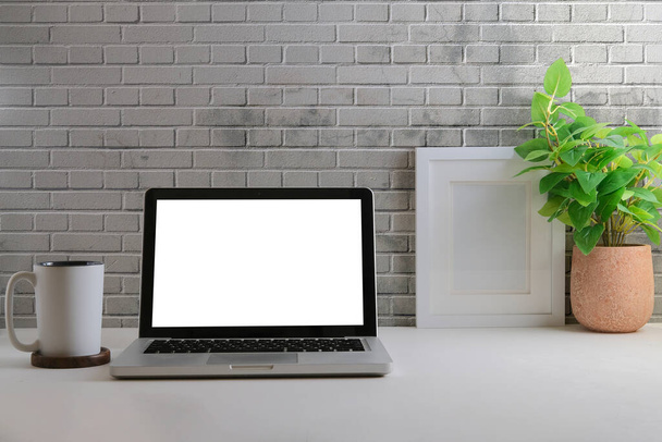 Home Office Schreibtisch, Laptop, Kaffeetasse, Topfpflanze und Bilderrahmen auf weißem Tisch mit Ziegelwand. - Foto, Bild