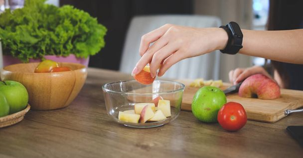 Woman cutting apple on wooden board, preparing fruit salad in the kitchen.	 - Foto, afbeelding