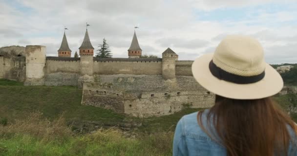 Achteraanzicht, vrouw met lang haar in een hoed, kijkt naar het oude kasteel fort. Overdag, middelmatig schot, bewolkt. Kamenets Podolsky, Oekraïne. - Video