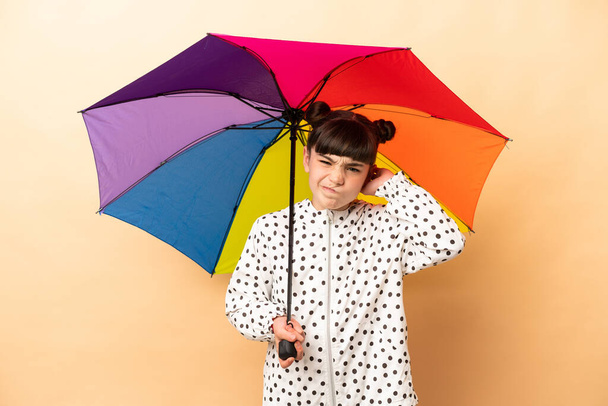 Little girl holding an umbrella isolated on beige background having doubts - Photo, Image