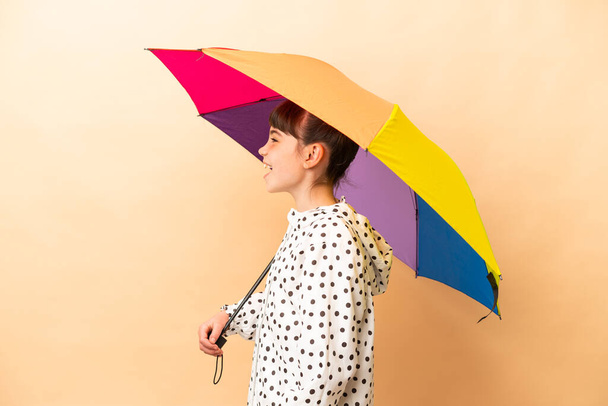 Mała dziewczynka trzyma parasol odizolowany na beżowym tle śmiejąc się w pozycji bocznej - Zdjęcie, obraz