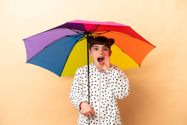 Mała dziewczynka trzyma parasol odizolowany na beżowym tle z zaskoczeniem i wstrząśniętym wyrazem twarzy - Zdjęcie, obraz
