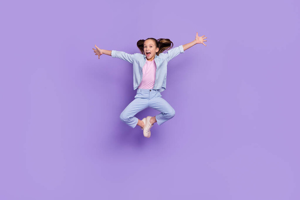 Photo of pretty funky little school girl wear denim shirt arms sides jumping high isolated purple color background - Foto, afbeelding