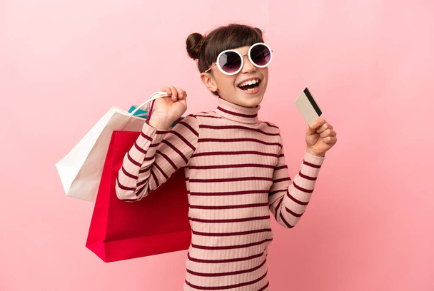 Little caucasian girl isolated on pink background holding shopping bags and a credit card - Фото, изображение