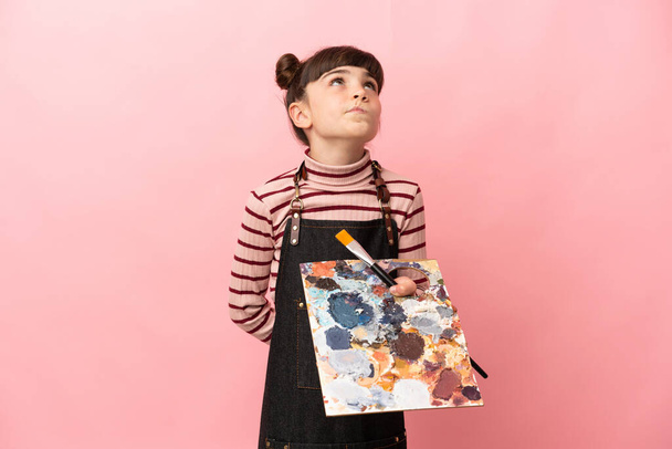 Little artist girl holding a palette isolated on pink background and looking up - Foto, imagen