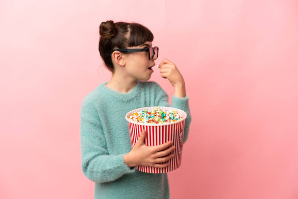Little caucasian girl isolated on pink background with 3d glasses and holding a big bucket of popcorns while looking side - Photo, Image