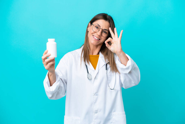 Жінка середнього віку ізольована на синьому фоні в одязі лікаря і тримає таблетки
 - Фото, зображення