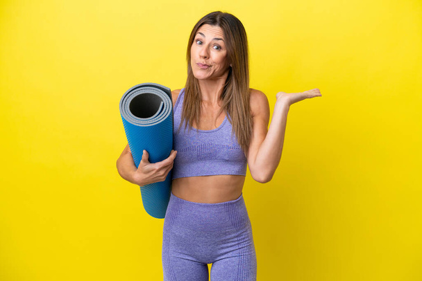 Femme sportive allant à des cours de yoga tout en tenant un tapis isolé fond non jaune ayant des doutes tout en levant la main - Photo, image